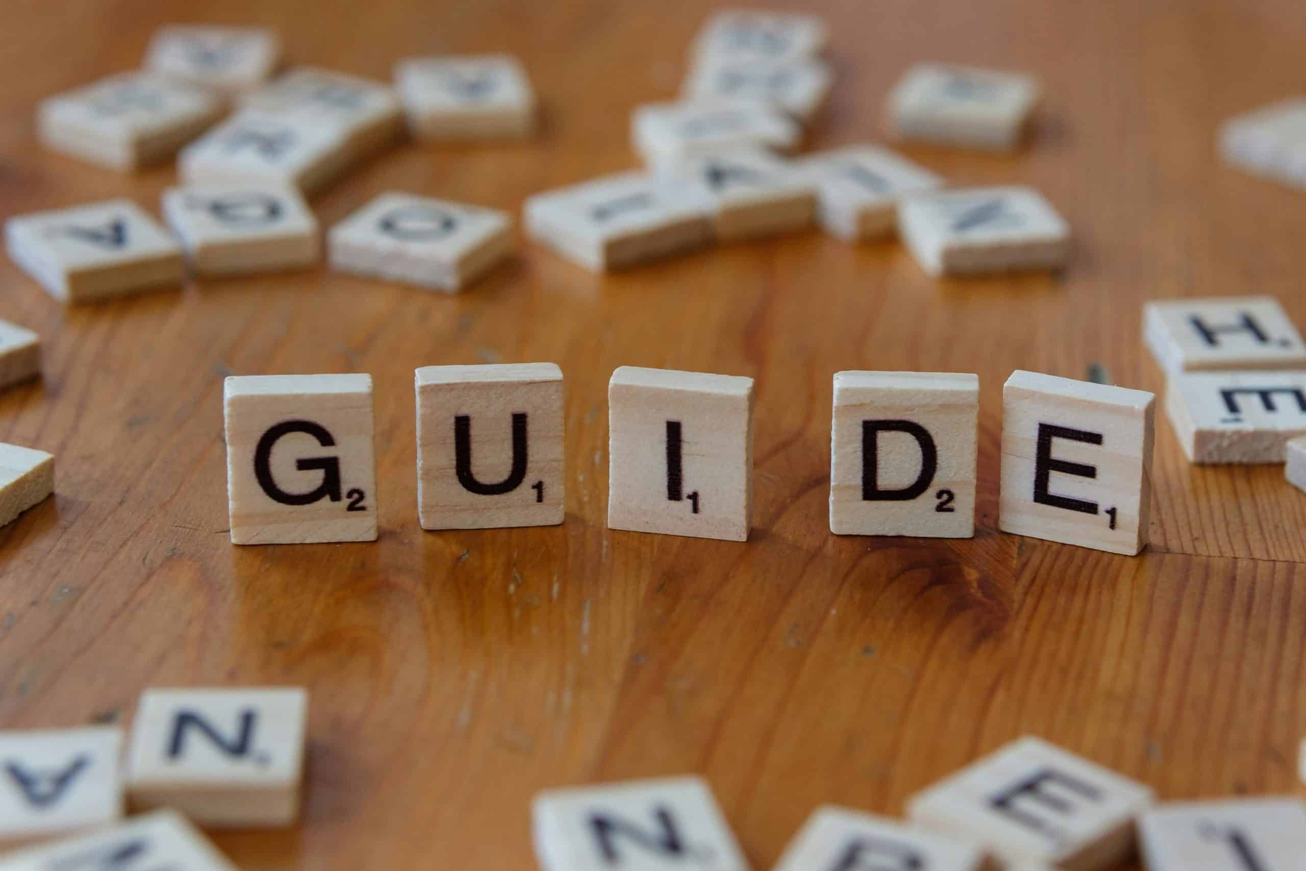Scrabble tiles forming the word 'Guide' surrounded by scattered letters on a wooden table.